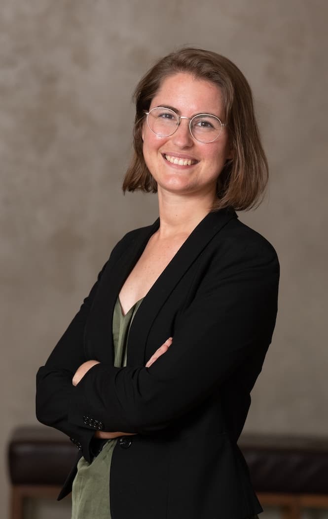 Siobhan stands with her arms crossed in front of a grey background. She is wearing a black blazer and a green blouse. Her hair is cropped just below her chin. She has a large smile.