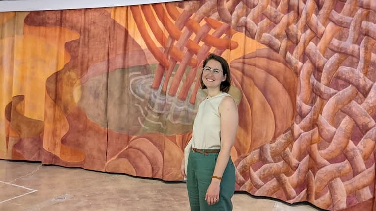 Siobhan stands in front of an oversized orange and peach canvas mural with abstract designs. She is wearing a cream blouse and green pants. She is smiling.