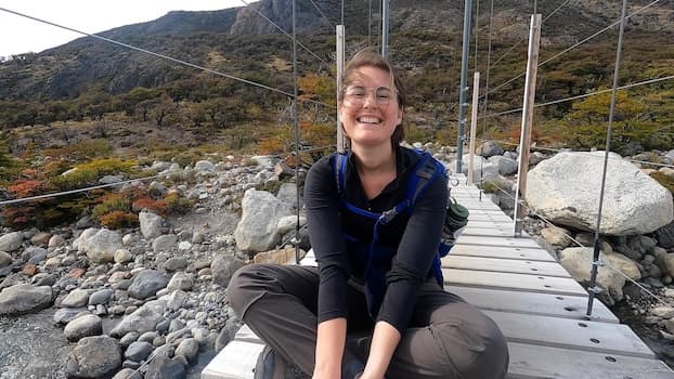 Siobhan is sitting cross legged with her hands by her feet. She is wearing grey and black hiking clothing and a blue back pack. She is mid laugh with her hair blowing in her face. She is sitting on a wooden bridge over a rocky river. There is lush greenery visible in the background.