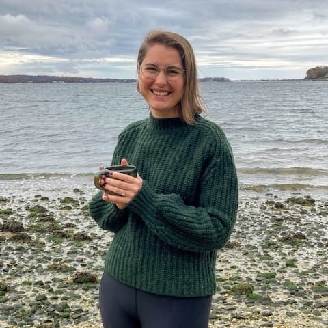 Siobhan is a light skinned woman with shoulder length brown hair. She wears thin framed glasses and is smiling bright. She stands on a rocky beach on an overcast day holding a mug in both hands. She is wearing a large green cozy sweater and black leggings.